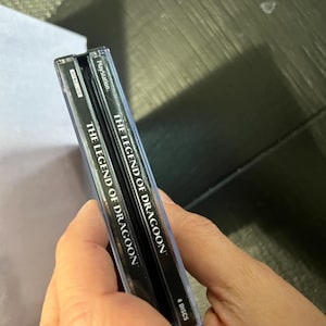 K&ouml;nnte beinhalten: Zwei schwarze PlayStation-Spieleboxen mit silbernem Text. Der Text auf den Boxen lautet "The Legend of Dragoon" und "4 Discs".