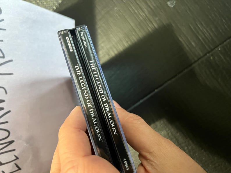 K&ouml;nnte beinhalten: Zwei schwarze PlayStation-Spieleboxen mit silbernem Text. Der Text auf den Boxen lautet "The Legend of Dragoon" und "4 Discs".