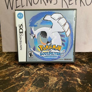 May include: A silver Nintendo DS game cartridge for Pokemon SoulSilver Version. The game features a white and blue background with a white Pokemon on it. The ESRB rating is E for Everyone. The text "The Pokemon Company" is visible on the cartridge.