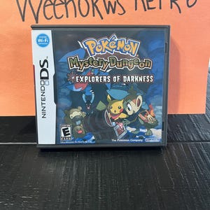 May include: A Nintendo DS game cartridge for Pokemon Mystery Dungeon: Explorers of Darkness. The game features a blue, black, and white design with images of Pikachu, a Grovyle, and a Squirtle. The game is rated E for Everyone by the PEGI.