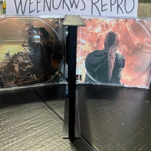 K&ouml;nnte beinhalten: Zwei transparente CD-H&uuml;llen aus Kunststoff mit schwarzen R&uuml;ckgraten, jede mit einer CD mit einem farbenfrohen Bild. Die linke CD zeigt ein dunkles Bild einer Person in einer dunklen Umgebung, w&auml;hrend die rechte CD ein helles Bild einer Person in einer feurigen Umgebung zeigt. Der Text "WEENORWS REPRO" ist auf einem wei&szlig;en Papier hinter den H&uuml;llen sichtbar.
