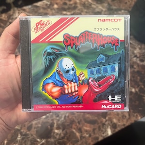 Op de afbeelding: Een zwarte en rode videospelcartridge voor het PC Engine-systeem. De game heet "Splatterhouse" en toont een monster met een grote bek en een man met een hockeymasker. De tekst op de cartridge luidt "©1988 1990 NAMCO LTD.. ALL RIGHTS RESERVED" en "namcoT スプラッターハウス HE System HuCARD".
