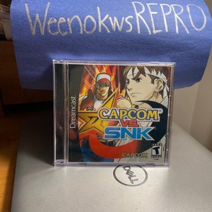 May include: A Dreamcast video game case with a red, yellow, and blue design. The game is called "CAPCOM VS. SNK" and features a red-haired character with a white headband and a blue-haired character with a white headband. The game is rated T for Teen by the ESRB.