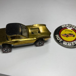 May include: A gold Hot Wheels toy car with a black roof and a black and yellow Hot Wheels button with the text "Mattel Python Hot Wheels" and "Original Mattel Inc. Hong Kong".