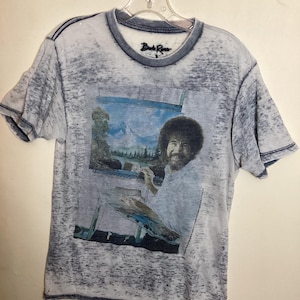 May include: A light blue t-shirt featuring a portrait of Bob Ross painting a landscape. The image is printed on the front of the shirt. The shirt has a distressed look with a dark blue trim around the collar and sleeves. The text "Bob Ross" is printed on the collar.