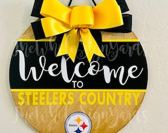 Könnte beinhalten: Ein runder Holztürhänger mit einem schwarz-gelben Steelers-Logo, den Worten "Willkommen im Steelers-Land" und einer großen gelben Schleife.