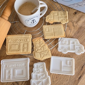 Könnte beinhalten: Ein Set mit sechs Ausstechern in Form eines Postamtes, eines Stapels Umschläge, eines Postwagens und einer Postkarte. Die Ausstecher sind aus weißem Kunststoff und befinden sich auf einer Holzoberfläche.