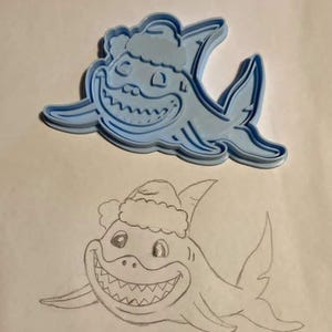 Könnte beinhalten: Ein hellblauer Ausstecher in Form eines Cartoon-Hais mit Weihnachtsmütze. Darunter befindet sich eine Bleistiftskizze des gleichen Haidesigns. Der Hai hat ein breites, zahnreiches Grinsen und Flossen.