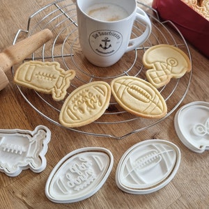 Könnte beinhalten: Ein Set mit sechs weißen Plastik-Ausstechern in Form von Fußbällen und Helmen. Die Ausstecher liegen auf einer Holzoberfläche mit einem Metall-Abkühlgitter und einer Tasse Kaffee.