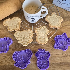 Könnte beinhalten: Ein Set mit neun Ausstechern in Form von Koalabären in verschiedenen Halloween-Kostümen. Die Ausstecher sind aus lila Kunststoff und auf einer Holzoberfläche angeordnet.