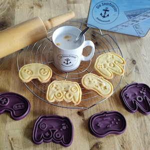 Könnte beinhalten: Lila und gelbe Ausstecher in Form von Videospielcontrollern. Die Ausstecher liegen auf einer Holzoberfläche mit einer Tasse Kaffee und einem Nudelholz.