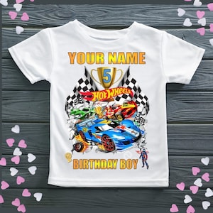 May include: White t-shirt with a Hot Wheels design. It features race cars, checkered flags, and the text "YOUR NAME" and "BIRTHDAY BOY". A gold trophy displays the number 5. The shirt is designed for a child.