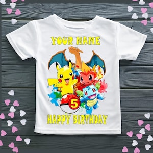 May include: White t-shirt with a colourful graphic of cartoon characters, a red and white ball with the number 5, and the text "YOUR NAME" and "HAPPY BIRTHDAY" in yellow. The design includes yellow, red, blue, and orange characters.