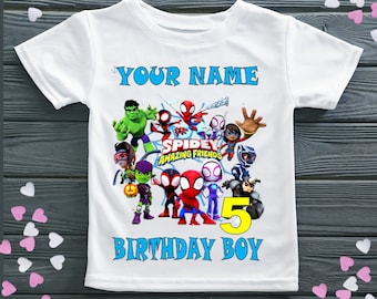 Könnte beinhalten: Weißes T-Shirt mit einem blauen "YOUR NAME"-Text oben. Das Shirt zeigt eine bunte Grafik von Spider-Man und seinen Freunden aus der Show "Spidey and his Amazing Friends". Die Grafik enthält das Logo der Show und die Zahl "5" in Gelb. Der Text "BIRTHDAY BOY" ist in Blau am unteren Rand des Shirts.