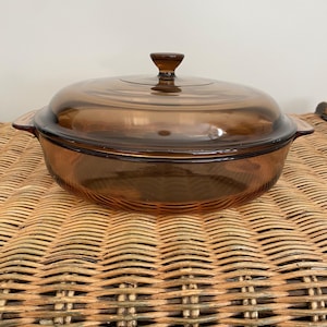 Può includere: Una pirofila vintage in vetro color ambra con coperchio abbinato. Il piatto ha una forma arrotondata, con due manici laterali e un pomello sul coperchio. Il vetro trasparente permette di vedere il contenuto.