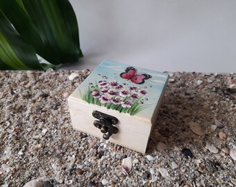 Puede incluir: Una caja cuadrada de madera con una escena pintada a mano en la tapa. La ilustración muestra una mariposa rosa y negra sobre un grupo de flores moradas y blancas con tallos verdes. La caja tiene un cierre metálico de color bronce.