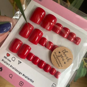 May include: A set of glossy, square-shaped, bright red press-on toenails in a clear plastic package. The packaging features social media icons and the hashtags #nailart, #naildesign, and #nailartist. A small sticker says "Thank you for supporting my small business."