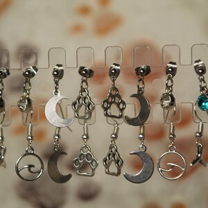 Puede incluir: Un conjunto de pendientes de plata con diferentes encantos, incluyendo una luna creciente, una huella de pata, una ola y un pez. Los pendientes se muestran en un soporte de plástico transparente.