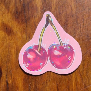 May include: A pink sticker with a cartoon illustration of two red cherries with green stems and leaves. The cherries have a glittery finish.
