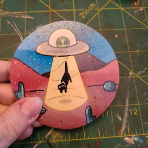 May include: A round sticker with a black cat being abducted by a UFO. The UFO is flying over a pink and blue desert landscape with cacti and stars. The sticker has a glittery finish.
