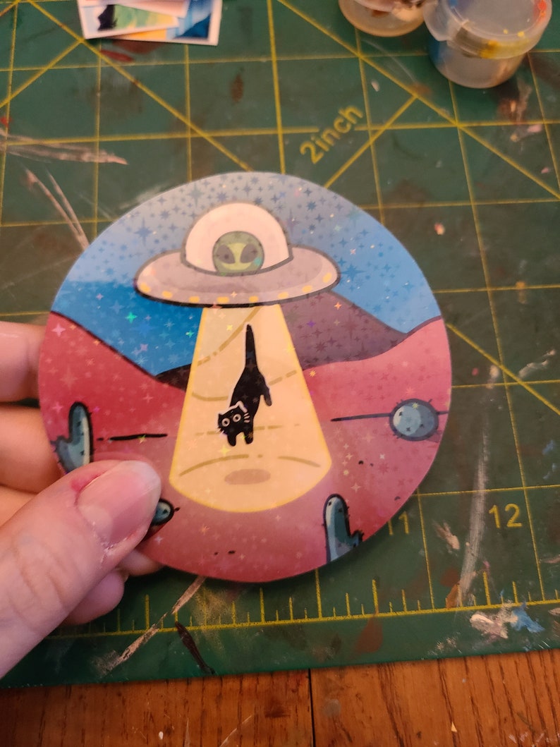 May include: A round sticker with a black cat being abducted by a UFO. The UFO is flying over a pink and blue desert landscape with cacti and stars. The sticker has a glittery finish.