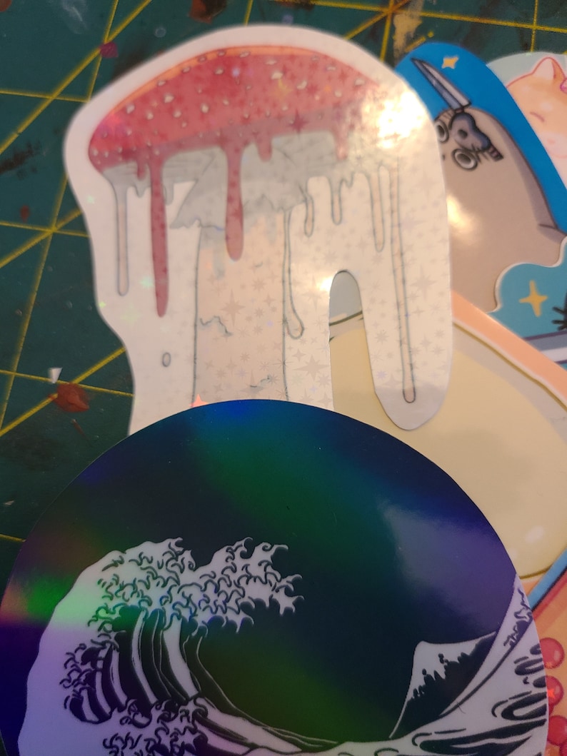 K&ouml;nnte beinhalten: Ein holografischer Aufkleber eines gro&szlig;en, tropfenden Pilzes mit einem rosa-wei&szlig;en Design. Der Aufkleber befindet sich auf einer gr&uuml;n-gelben Oberfl&auml;che.