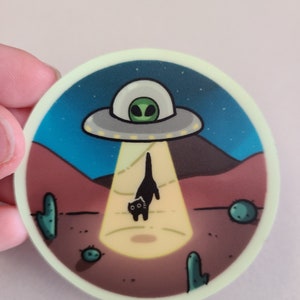 May include: A black cat with green eyes walks away from a glowing UFO in a desert landscape with cacti. The UFO is hovering above the cat and casting a beam of light on the ground.