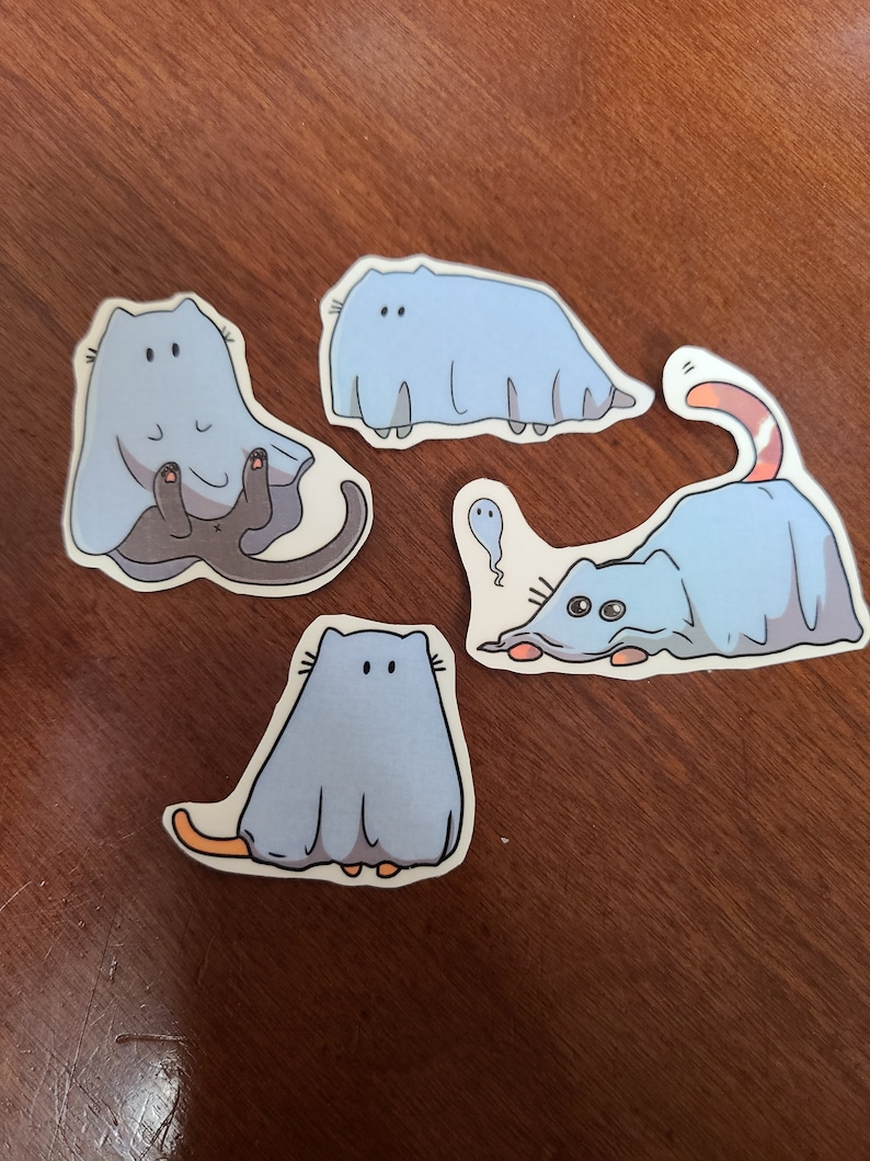 May include: Four stickers featuring cartoon cats dressed as ghosts. The cats are in various poses, with some peeking out from under the sheet and others fully covered. The stickers are on a wooden surface.