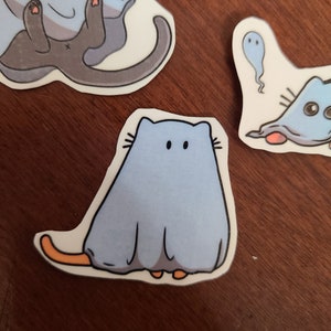 May include: A blue and white ghost cat sticker with orange feet and whiskers. The cat is wearing a ghost costume with a white sheet and a blue face.