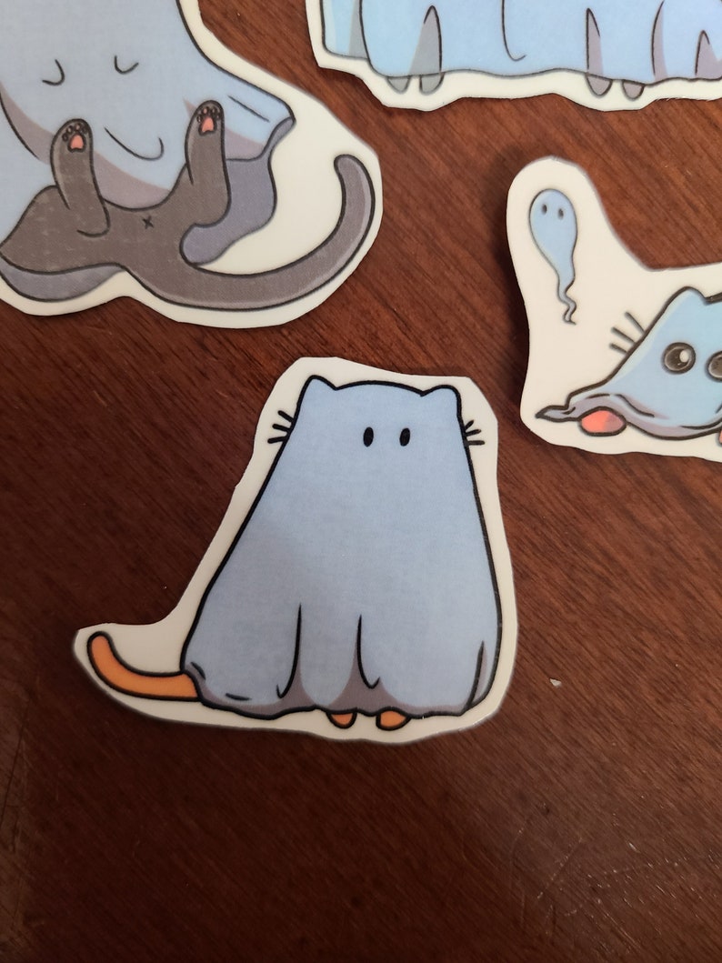 May include: A blue and white ghost cat sticker with orange feet and whiskers. The cat is wearing a ghost costume with a white sheet and a blue face.