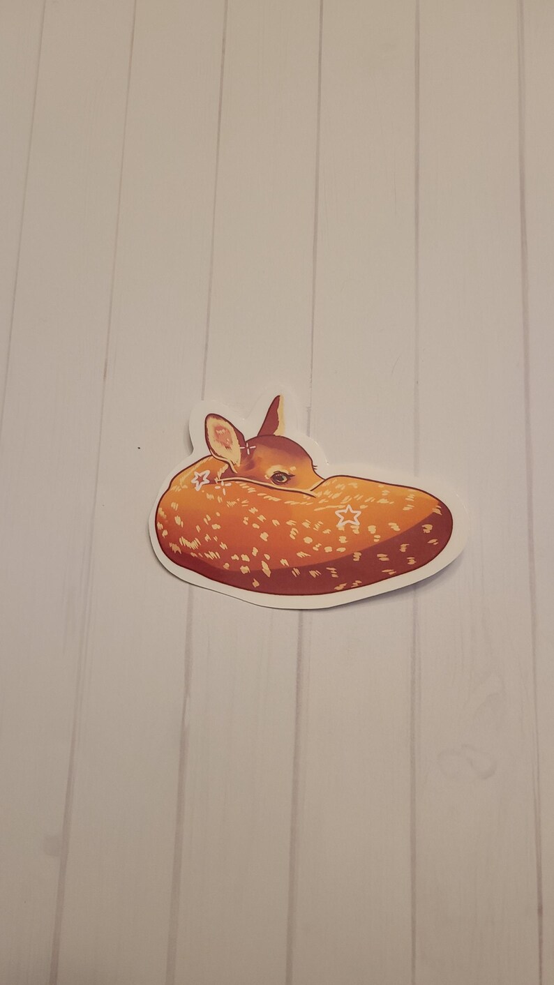 May include: A sticker featuring a sleeping fawn. The fawn is curled up, with its head tucked in. The sticker has a white border and is primarily orange and brown with white star accents.