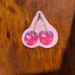 May include: A sticker of two red cherries with green stems and leaves on a pink background with glitter accents.