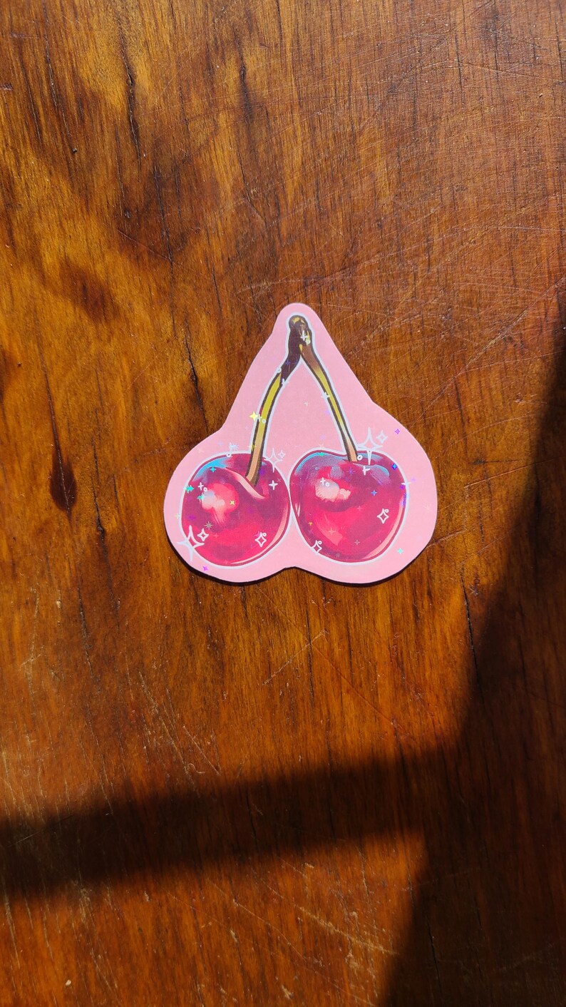 May include: A sticker of two red cherries with green stems and leaves on a pink background with glitter accents.