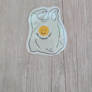 May include: A sticker in the shape of a plastic shopping bag with the words "NO THANK YOU" and a smiling face. The sticker is white with black outlines and is on a wooden surface.