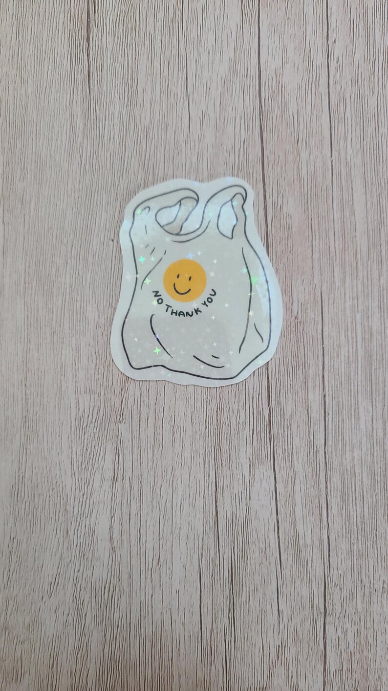 May include: A sticker in the shape of a plastic shopping bag with the words "NO THANK YOU" and a smiling face. The sticker is white with black outlines and is on a wooden surface.