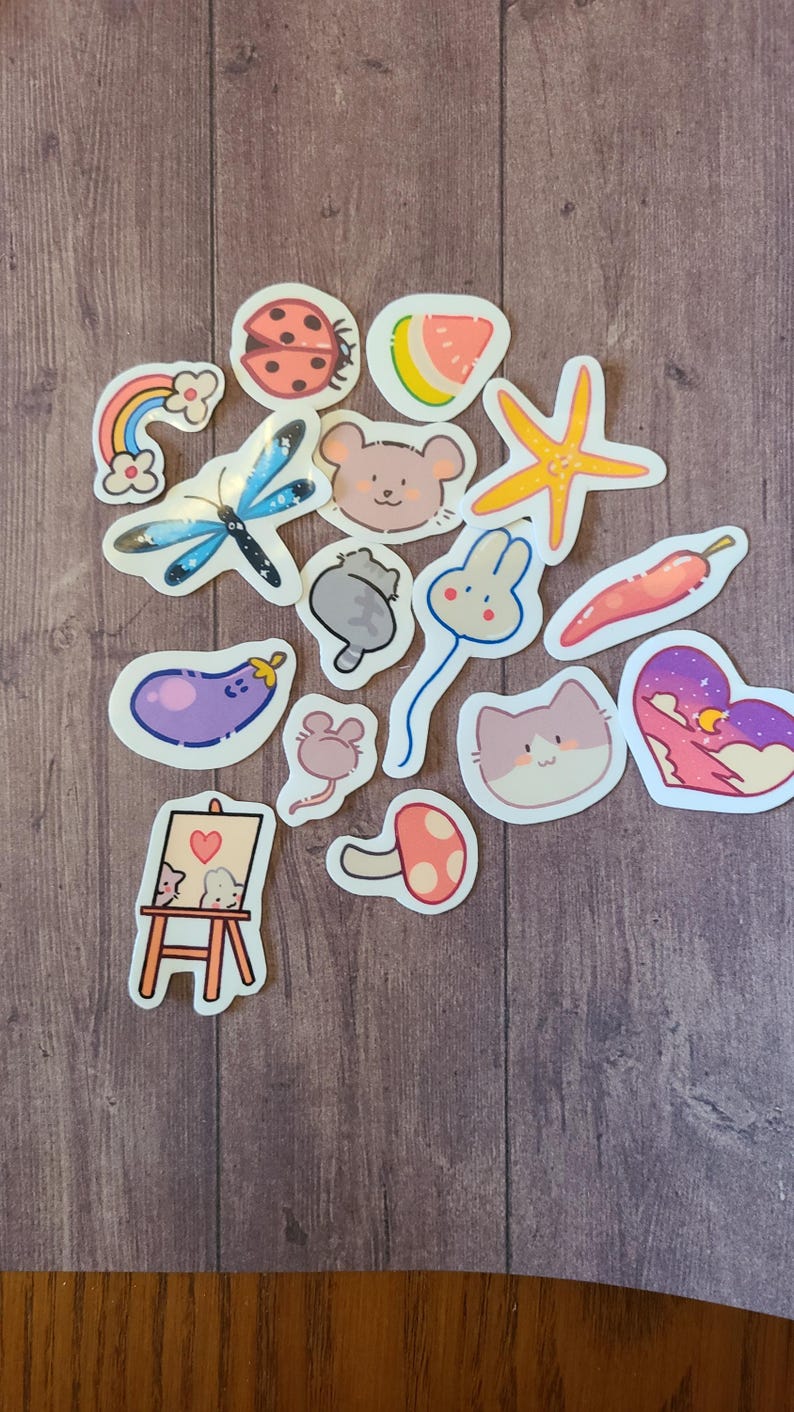 Op de afbeelding: Een verzameling kleurrijke stickers met verschillende schattige dier- en natuurontwerpen. De stickers bevatten een lieveheersbeestje, een watermeloen, een zeester, een vlinder, een muis, een konijn, een kat, een aubergine, een paddenstoel en een regenboog.