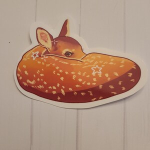 May include: A sticker featuring a curled-up fawn with a light brown and white spotted coat. The fawn's head is visible, with its ears and eyes showing. The sticker has a white border and is set against a light wood-grain background.
