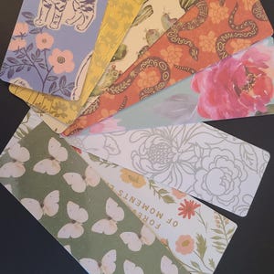 May include: A set of eight colorful bookmarks featuring various designs, including butterflies, flowers, cats, snakes, and cacti. The bookmarks are all rectangular and have a white background.
