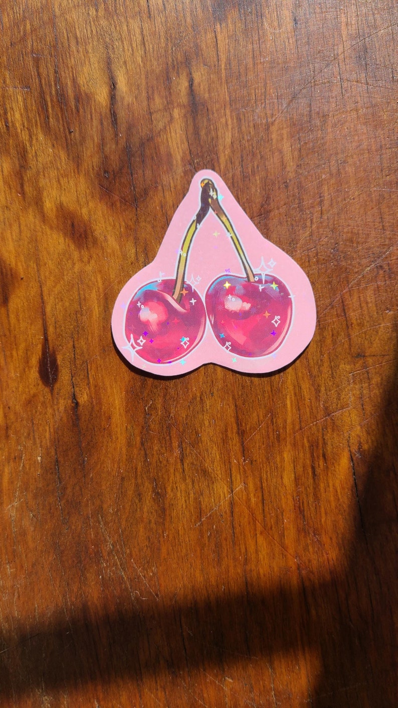 May include: A pink sticker with a cartoon illustration of two red cherries with green stems and leaves. The cherries have a glittery finish.