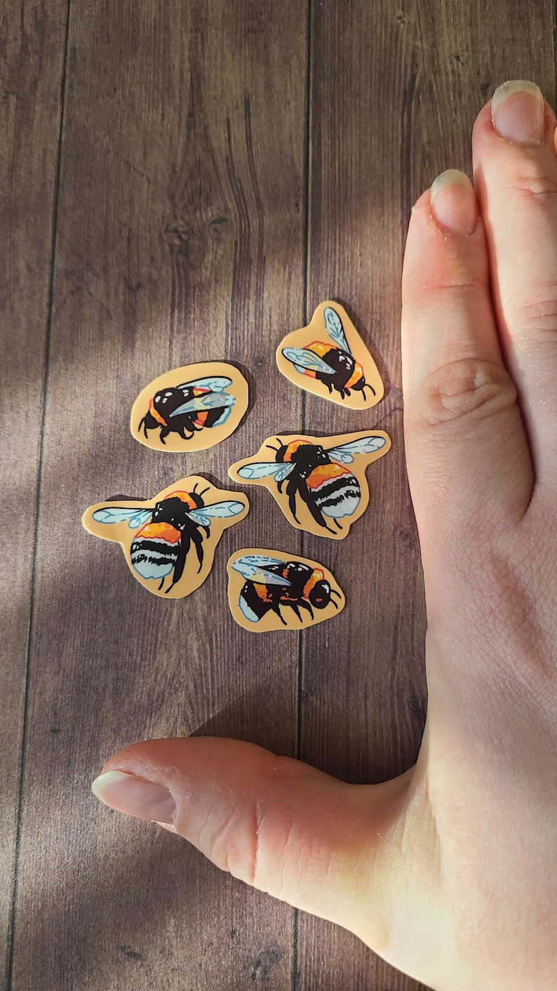 Op de afbeelding: Vijf gele en zwarte hommelstickers op een houten oppervlak. De hommels bevinden zich in verschillende posities, sommige vliegen en andere rusten.