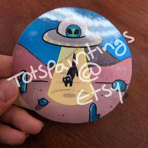 May include: A round sticker with a black cat walking in front of a UFO. The UFO is hovering over a desert landscape with cacti and a yellow beam of light shining down. The text "Totspantings @ Etsy" is written on the sticker.