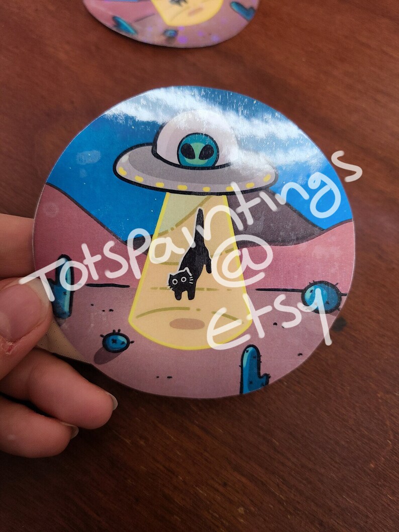 May include: A round sticker with a black cat walking in front of a UFO. The UFO is hovering over a desert landscape with cacti and a yellow beam of light shining down. The text "Totspantings @ Etsy" is written on the sticker.