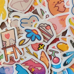 Op de afbeelding: Een verzameling kleurrijke stickers met verschillende schattige illustraties, waaronder een regenboog, een beer die een hart vasthoudt, een bloem, een taart, een konijn, een kat, een paddenstoel, een wolk, een zon, een origami-kraanvogel en een schildersezel.