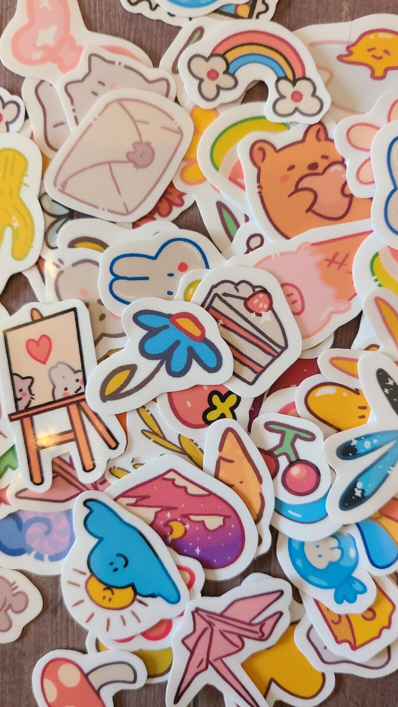 Op de afbeelding: Een verzameling kleurrijke stickers met verschillende schattige illustraties, waaronder een regenboog, een beer die een hart vasthoudt, een bloem, een taart, een konijn, een kat, een paddenstoel, een wolk, een zon, een origami-kraanvogel en een schildersezel.