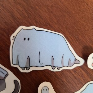 May include: A light blue sticker of a cartoon cat with a long, thin body and two large eyes. The cat is wearing a white ghost costume.