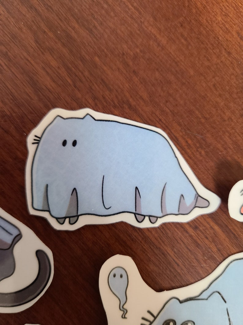 May include: A light blue sticker of a cartoon cat with a long, thin body and two large eyes. The cat is wearing a white ghost costume.