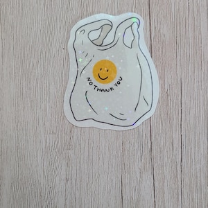 May include: A white sticker in the shape of a plastic shopping bag with the text "No thank you" and a yellow smiley face. The sticker has a holographic, glittery effect.
