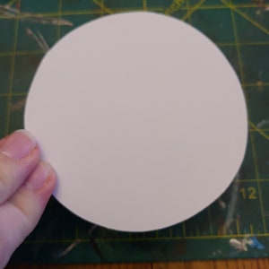 May include: A white, round, blank paper circle on a green cutting mat with yellow lines.