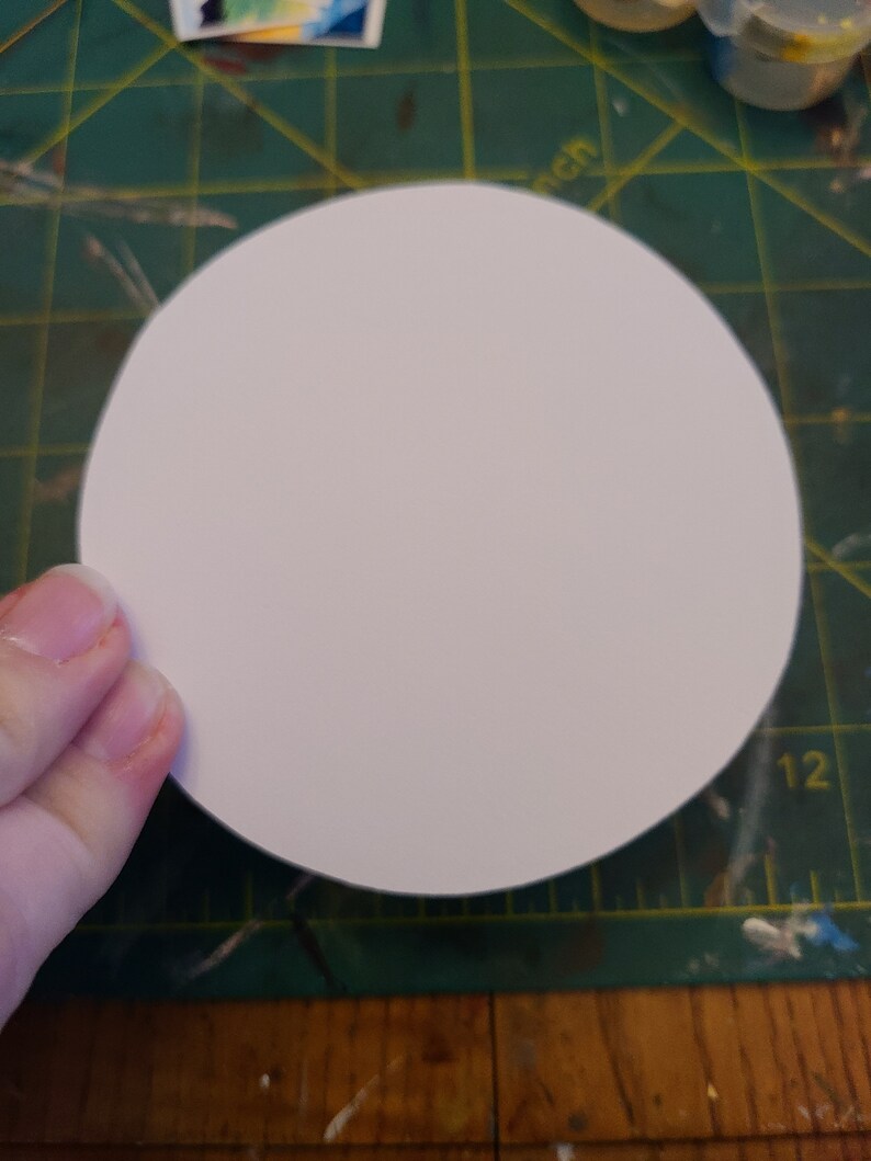 May include: A white, round, blank paper circle on a green cutting mat with yellow lines.