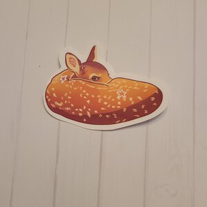May include: A sticker featuring a stylized illustration of a sleeping fawn. The fawn is depicted in shades of brown and orange with white spots and star accents. The sticker is on a white background.